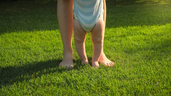 Primer plano del bebé descalzo y la madre de pie sobre hierba verde fresca en el parque. Concepto de estilo de vida saludable, desarrollo infantil y crianza. — Foto de Stock