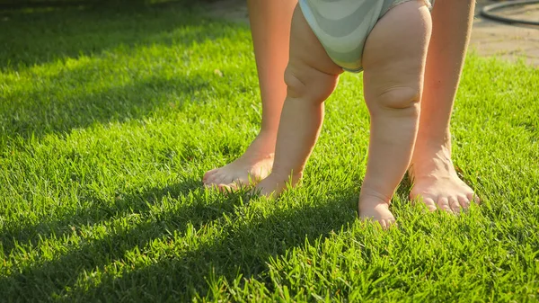 新鮮な緑の芝生の芝生の上で最初の一歩を踏み出す彼女の赤ちゃんの息子をサポートする母親の閉鎖。健康的なライフスタイル、子供の発達と子育ての概念. — ストック写真
