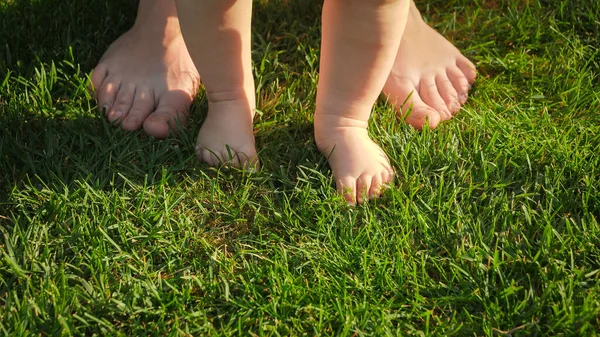 Close-up de bebê descalço em pé na grama verde fresca com a mãe. Conceito de estilo de vida saudável, desenvolvimento infantil e parentalidade. — Fotografia de Stock