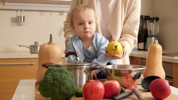 Очаровательный улыбающийся мальчик сидит на кухонном столе, полном овощей, фруктов и посуды. Концепция маленького шеф-повара, детская кухня, хорошее семейное время вместе — стоковое видео
