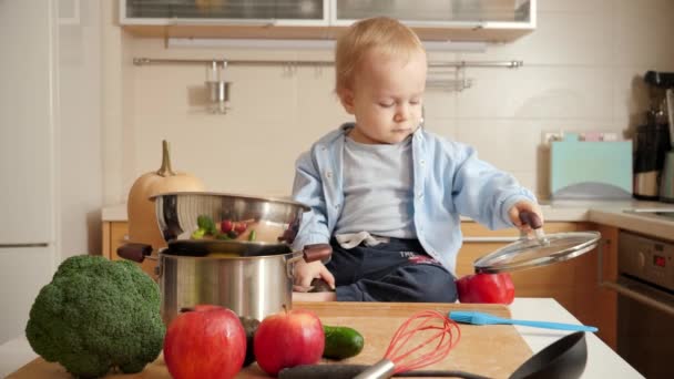 Schattig jongetje dat speelt met metalen pannen, glazen deksels en kommetje op de keuken. Concept van kleine chef-kok, kinderen koken voedsel — Stockvideo