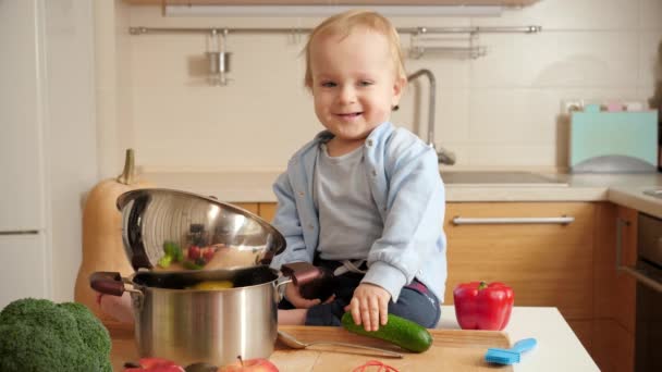 Schattig lachend jongetje zittend op houten keukentafel en spelend met groenten. Concept van kleine chef-kok, kinderen koken voedsel — Stockvideo