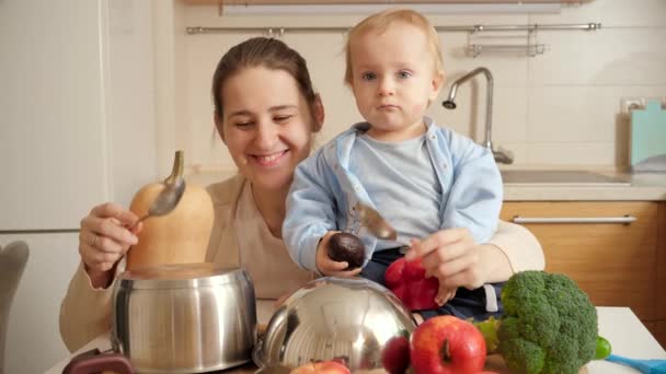 Χαρούμενη χαμογελαστή μητέρα με τον μικρό γιο να παίζει με κουτάλια σε τηγάνια στην κουζίνα όπως στα τύμπανα. Έννοια του μικρού σεφ, τα παιδιά μαγείρεμα των τροφίμων, καλές οικογενειακές στιγμές μαζί. — Αρχείο Βίντεο