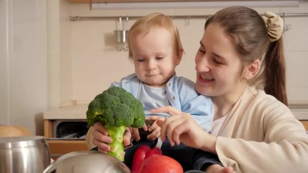 Sonriente joven madre mostrando brócoli a su hijo bebé mientras cocina en la cocina. Concepto de pequeño chef, niños cocinando comida, buen tiempo en familia juntos. — Vídeo de stock