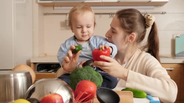 Glimlachende moeder met baby jongen die liedjes speelt en zingt met groenten en fruit in de keuken. Concept van kleine chef-kok, kinderen koken eten, goede familie tijd samen. — Stockvideo