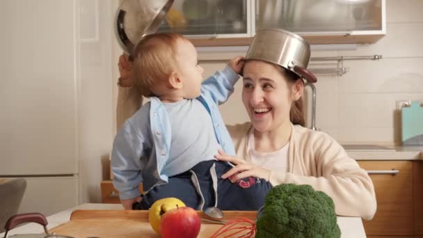 Gelukkige lachende moeder met zoontje spelend in de keuken met pannen op het hoofd. Concept van kleine chef-kok, kinderen koken eten, goede familie tijd samen. — Stockvideo