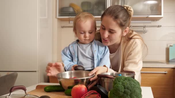Νεαρή μητέρα δείχνει μαγειρικά σκεύη κουζίνας, σκεύη και συστατικά στο μικρό γιο της στην κουζίνα. Έννοια του μικρού σεφ, τα παιδιά μαγείρεμα των τροφίμων, καλές οικογενειακές στιγμές μαζί. — Αρχείο Βίντεο
