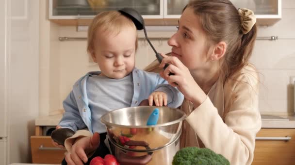 Χαρούμενη χαμογελαστή μητέρα που παίζει με την κουτάλα και τον μικρό της γιο ενώ μαγειρεύει στην κουζίνα. Έννοια του μικρού σεφ, τα παιδιά μαγείρεμα των τροφίμων, καλές οικογενειακές στιγμές μαζί. — Αρχείο Βίντεο