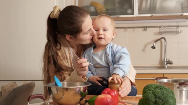 Усміхнена молода мати годує свого маленького сина, сидячи на кухонному столі. Концепція маленького шеф-кухаря, дітей, які готують їжу, гарний сімейний час разом — стокове відео