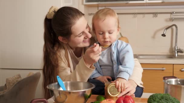 台所のテーブルに座って調理器具で遊んでいるかわいい赤ちゃんの男の子とお粥で彼を養う母親。小さなシェフのコンセプト、子供たちが料理を作る、楽しい家族時間を一緒に. — ストック動画