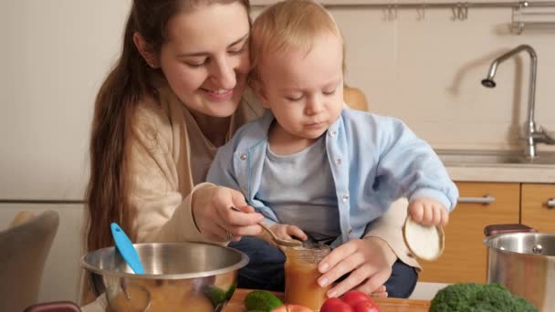 Усміхнена жінка дає фруктове пюре своєму маленькому синові, який грає на кухні. Концепція маленького шеф-кухаря, дітей, які готують їжу, гарний сімейний час разом . — стокове відео