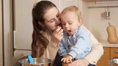 Gülümseyen anne küçük bebeğini mutfak masasında meyve püresiyle besliyor. Küçük şef kavramı, çocukların yemek pişirmesi, sağlıklı beslenme..