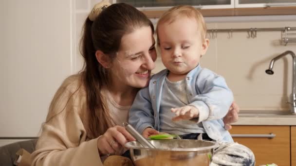 Feliz madre sonriente con su pequeño hijo jugando con utensilios de cocina mientras hace pan en la cocina. Concepto de pequeño chef, niños cocinando alimentos, nutrición saludable. — Vídeo de stock