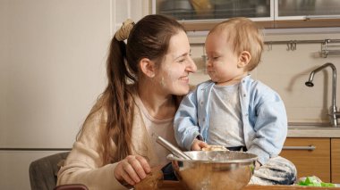 O evde ekmek pişirirken küçük oğlunu mutfak masasında kucaklayan mutlu bir anne. Küçük şef kavramı, çocukların yemek pişirmesi, sağlıklı beslenme..