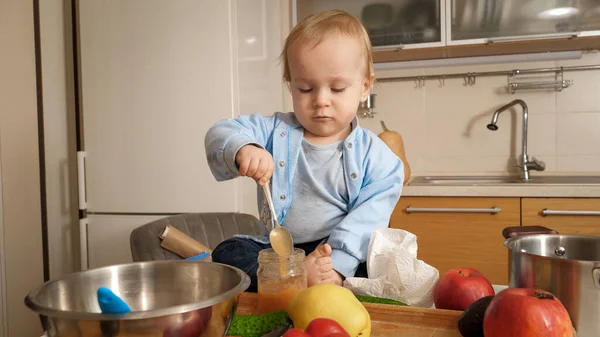 Piccolo bambino seduto sul tavolo della cucina e mangiare purea di frutta con cucchiaio. Concetto di alimentazione sana, dieta e cibo per bambini — Foto Stock