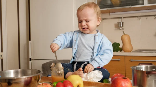Piccolo bambino che piange cercando di mangiare porridge con cucchiaio in cucina. Concetto di alimentazione sana, dieta e cibo per bambini — Foto Stock