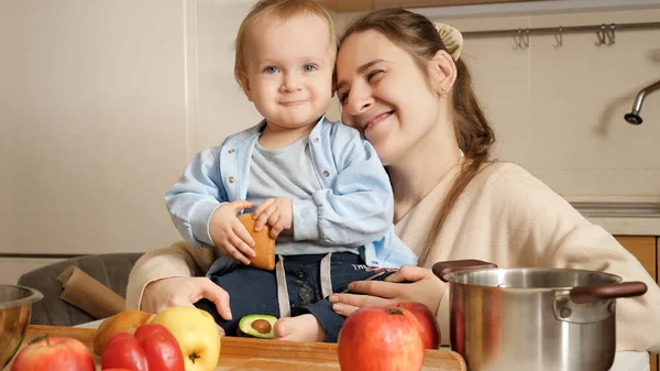 Šťastná usměvavá matka se dívá na svého malého syna, jak jí doma omastek v kuchyni. Koncepce zdravé výživy, stravy a dětské výživy. — Stock fotografie