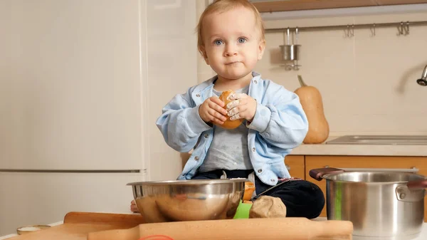 Portrét roztomilého usmívajícího se chlapečka, jak jí čerstvý chléb a hraje mouku na kuchyni. Koncept malého šéfkuchaře, děti vařící jídlo, zdravá výživa. — Stock fotografie