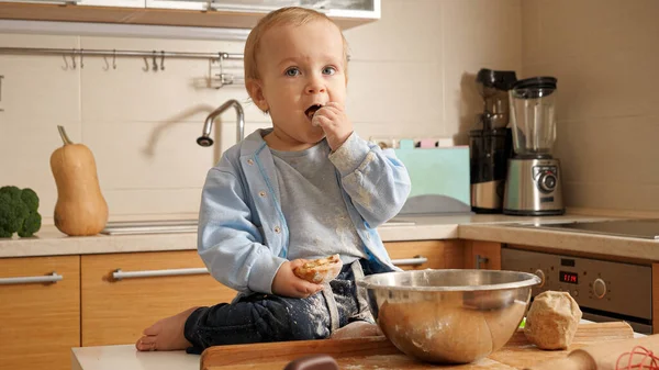 Küçük komik çocuk mutfakta ekmek hamuru yaparken un yiyor. — Stok fotoğraf