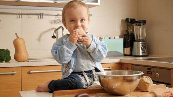 1 anno bambino coperto di farina e pasta mangiare pane appena sfornato in cucina — Foto Stock