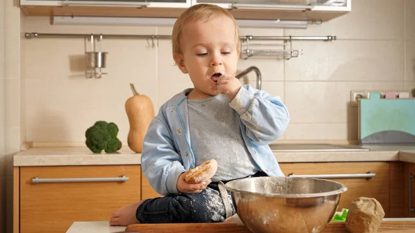 Słodki chłopiec siedzi na stole w kuchni, bawiąc się mąką i jedząc chleb. Pojęcie małego kucharza, dzieci gotujące jedzenie, zdrowe odżywianie. — Zdjęcie stockowe