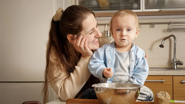 Gülümseyen anne, un ve hamura bulanmış bebeğine bakarken hamur yoğuruyor ve ekmek pişiriyor. — Stok fotoğraf