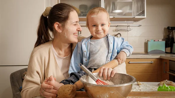 Счастливый улыбающийся мальчик с матерью, месящей тесто для выпечки хлеба на кухне. Концепция маленького шеф-повара, детская кухня, здоровое питание — стоковое фото