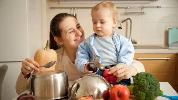 Felice madre sorridente con bambino che gioca con i cucchiai sulle padelle in cucina come alla batteria. Concetto di piccolo chef, cucina per bambini, buon tempo in famiglia insieme. — Foto Stock