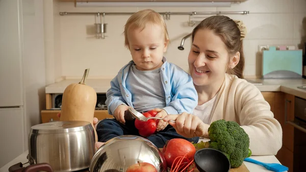 Sorridente giovane madre che mostra i broccoli al suo bambino mentre cucina in cucina. Concetto di piccolo chef, cucina per bambini, buon tempo in famiglia insieme. — Foto Stock