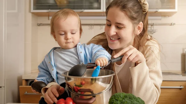 Felice madre sorridente che gioca con mestolo e il suo bambino mentre cucina in cucina. Concetto di piccolo chef, cucina per bambini, buon tempo in famiglia insieme. — Foto Stock