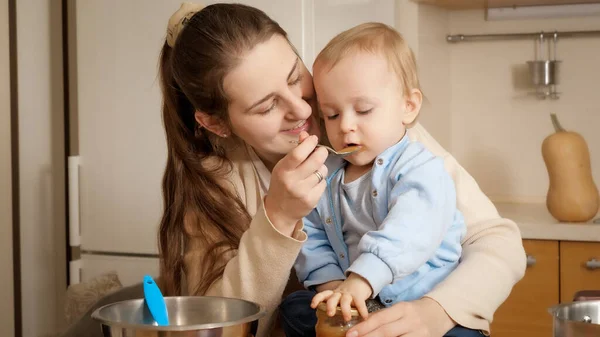 Usmívající se žena dává ovocné pyré svému malému synovi, který si hraje v kuchyni. Koncept malého šéfkuchaře, děti vařící jídlo, dobré společné rodinné chvíle. — Stock fotografie