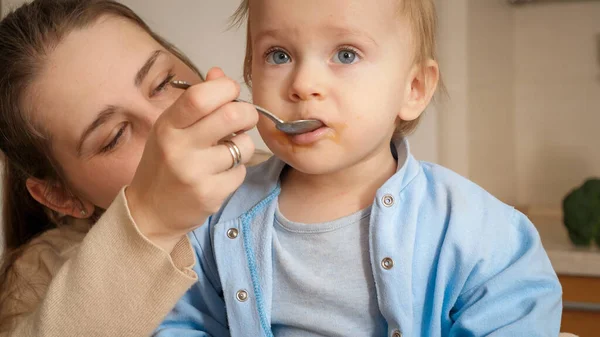 Closeup of little baby boy eating fruit purree on kitchen from spoon. Concept of little chef, children cooking food, healthy nutrition — Stock Photo, Image