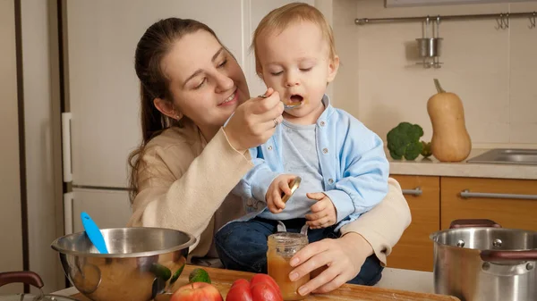 Giovane madre amorevole che alimenta il suo bambino che gioca con pentole in cucina. Concetto di piccolo chef, cibo per bambini, nutrizione sana — Foto Stock