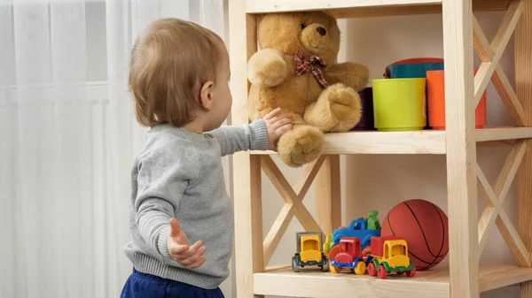 Cute baby boy taking teddy bear toy from bookshelf in playroom. Child education — Foto Stock