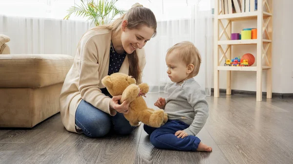 Happy smiling mother playing with her baby son with teddy bear on floor in living room. — Foto Stock