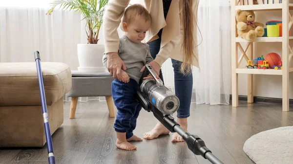 Young mother doing housework teaching and giving vacuum cleaner to her baby son. — Zdjęcie stockowe