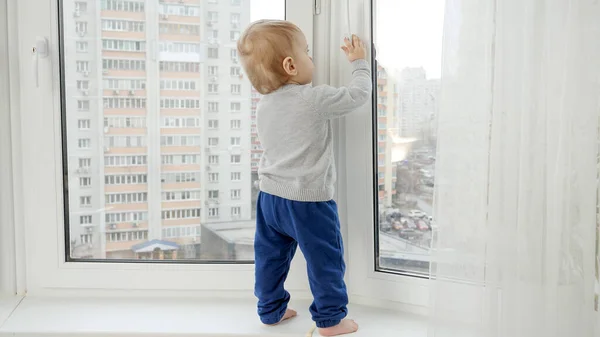 Piccolo bambino in piedi sul davanzale della finestra e tirando maniglia della finestra. Bambino in pericolo. Sicurezza dei bambini. — Foto Stock