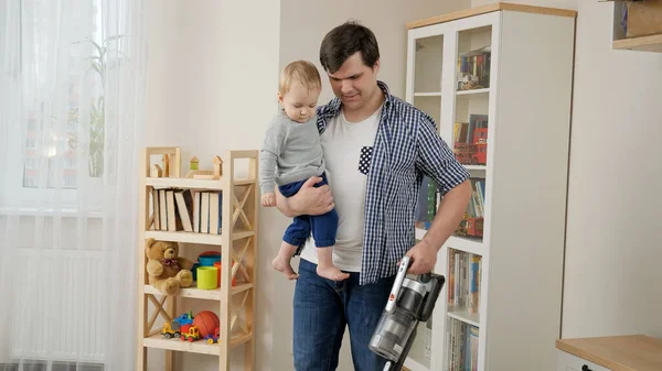 Jonge vader houdt zijn baby zoon terwijl het doen van huis opruimen en met behulp van stofzuiger. — Stockfoto