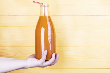 Sarı, ahşap arka plan. İçinde portakal suyu olan cam bir şişe. Mandalina ve havuç suyunun yanı sıra balkabağı da dişi bir el tarafından tutulur..