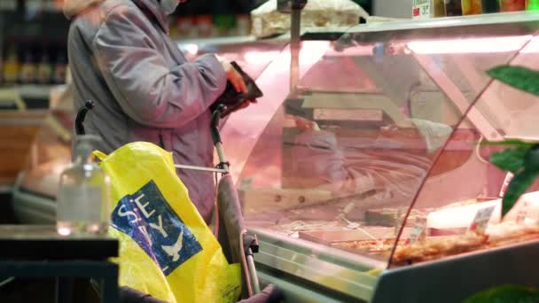 Une vieille dame achète de la viande dans un magasin. Un pensionné se tient devant une vitrine en verre — Video