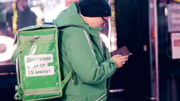 A food delivery courier in a green uniform with a thermal backpack checks orders — Stock Video