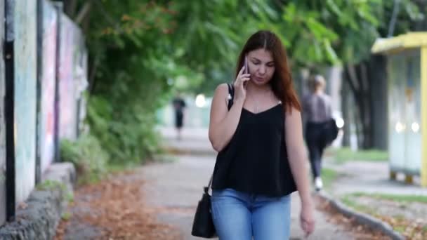 Giovane donna in jeans e camicetta sta parlando al telefono mentre cammina per strada — Video Stock