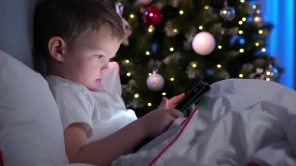 子供はラップトップでゲームをプレイし、リモートで学習します。小さなブロンド男の子にクリスマスの夜に背景にクリスマスツリー,ベッドに横たわってノートパソコンで彼の手と暗い部屋で漫画を見て. — ストック動画