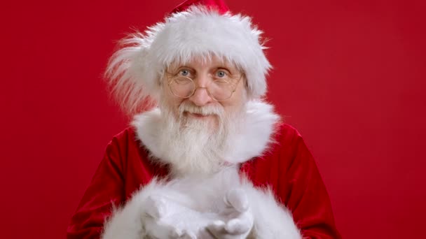サンタクロースの幸せな面白い肖像カメラを見て、赤の背景に立っている間に彼の手をオフに白い雪を吹いて。サンタクロースが手から雪を吹いている。クリスマスマジック. — ストック動画