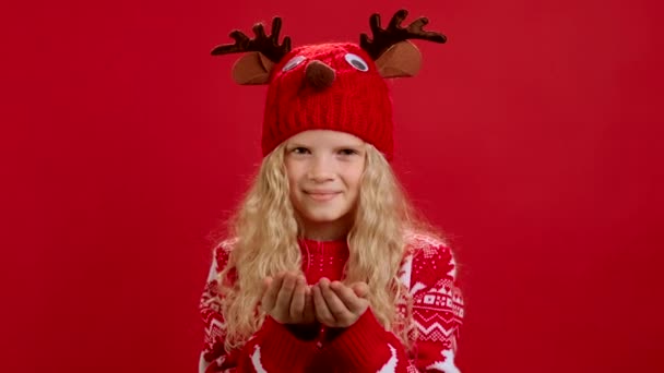 Portret Piękna blondynka w czerwonym swetrze i czerwonym kapeluszu z poroża jelenia, wieje biały puszysty śnieg z jej rąk, uśmiechy i fale Witam z rękami na czerwonym tle. Wesołych Świąt. — Wideo stockowe