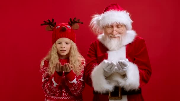 Neşeli Noel Baba ve Şenlikli Yeni Yıl Kıyafeti 'nde Genç Sarışın Kız Ellerindeki Beyaz Kar' ı Uçuruyor, Gülüyorlar ve Kırmızı Arkaplan 'da Ayakta Dururken Tezahürat Yapıyorlar. Noel Ruhu. — Stok video
