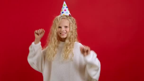 Щаслива дівчина в білому светрі і зі святковим конусом на голові танцює весело махаючи руками, святкуючи свій день народження. Блондинка підліток відзначає перемогу або велику перемогу танцює на червоному тлі . — стокове відео