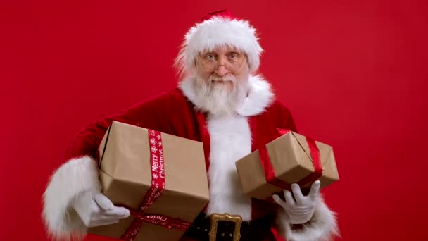 Funny Santa Claus en costume de Noël avec des boîtes-cadeaux sur fond rouge regarde la caméra et jette des boîtes de cadeaux en avant. Joyeux Père Noël jette des cadeaux. Joyeux Noël et cadeaux de Saint-Nicolas. — Video