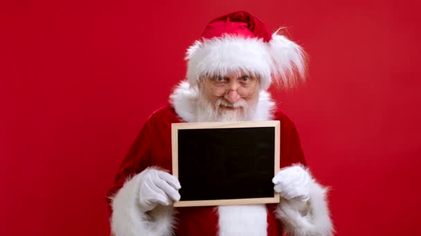 Kırmızı Kostümlü Portre Sakallı Noel Baba kameraya bakar ve çalışma alanı fotokopi mekanıyla elindeki siyah tabeladan dışarı bakar. İyi Noeller Alışveriş İçin Düşük Fiyatlar. — Stok video