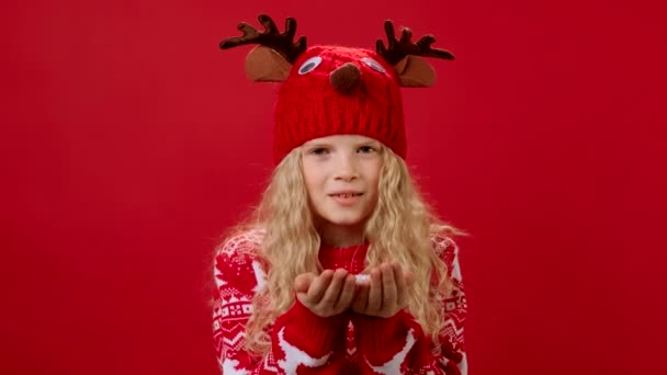 Portret Slow Motion Piękna blondynka w świątecznym czerwonym swetrze i czerwonym kapeluszu z poroża jelenia, wieje biały puszysty śnieg z jej rąk, uśmiechy i fale Witam z jej rąk na czerwonym tle. — Wideo stockowe